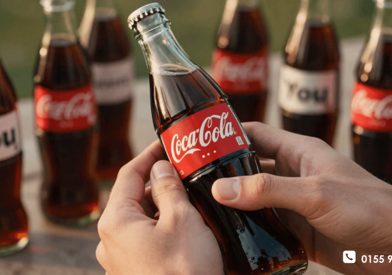 حملة شارك كوكاكولا .. كيف طبعت كولا أسماء الناس وزادت مبيعاتها؟ مشهد ملون ومبهج لمئات زجاجات كوكاكولا مصطفة، كل واحدة عليها اسم شخص مختلف مطبوع على الملصق، مع أيدي الناس تمدد لتختار أسمائهم، أجواء صيفية دافئة تعكس حملة "شارك كوكاكولا" الناجحة.